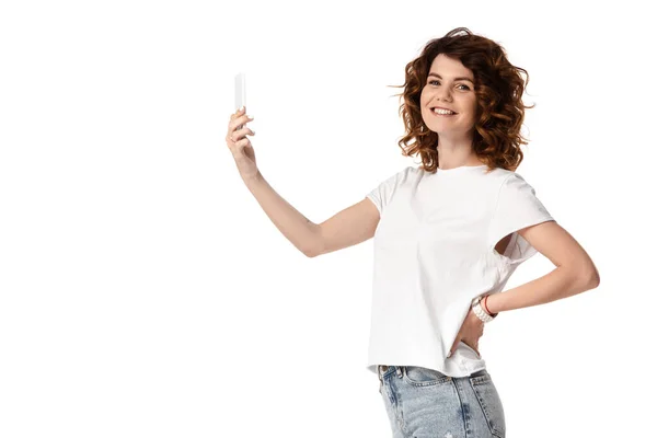 Mujer feliz sosteniendo el teléfono inteligente mientras está de pie con la mano en la cadera aislado en blanco - foto de stock