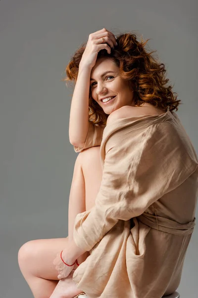 Curly woman smiling while sitting on chair isolated on grey — Stock Photo