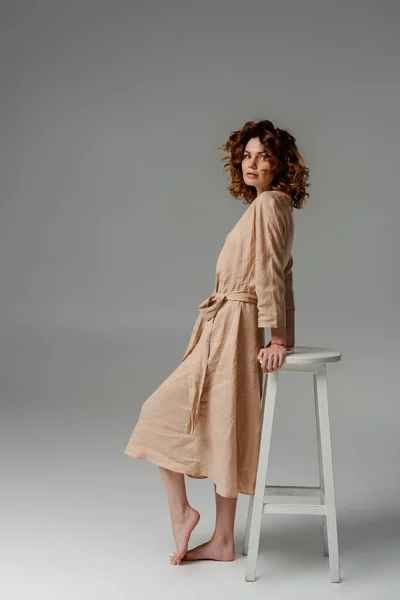 Femme bouclée et pieds nus debout près de la chaise sur gris — Photo de stock