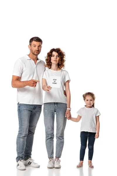 Handsome man pointing with finger at card with stay at home lettering near attractive wife and kid on white — Stock Photo