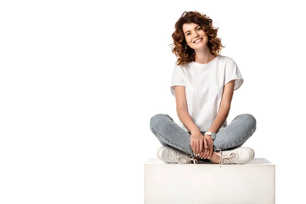 Весела жінка в джинсах сидить на кубі і посміхається ізольовано на білому — стокове фото