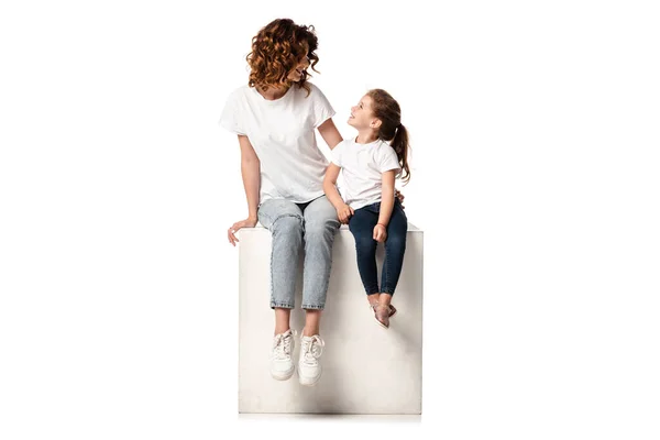 Happy mother and daughter looking at each other while sitting on cube on white — Stock Photo