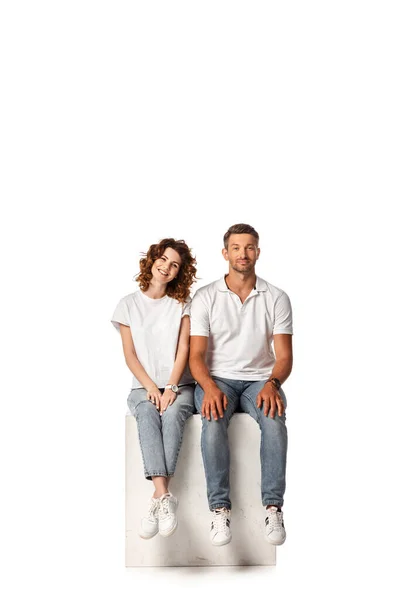 Cheerful couple smiling and sitting on cube on white — Stock Photo