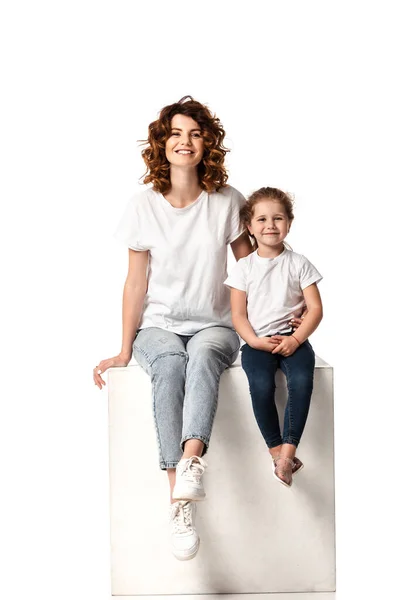 Happy mother and daughter looking at camera while sitting on cube on white — Stock Photo