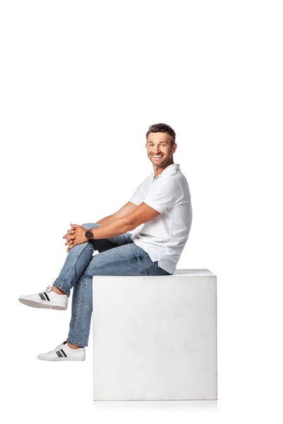 Happy man in denim jeans sitting on cube and looking at camera on white — Stock Photo