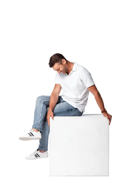 Felice uomo in jeans seduto sul cubo e guardando giù isolato su bianco — Foto stock