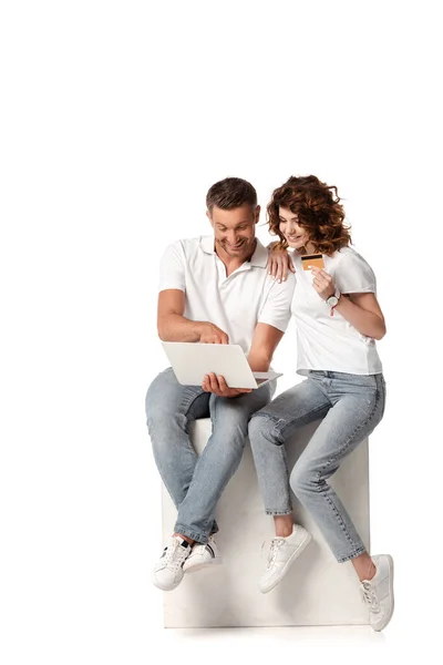 Mujer feliz sosteniendo la tarjeta de crédito cerca del hombre usando el ordenador portátil en blanco - foto de stock