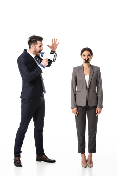 Hombre de negocios barbudo gritando en megáfono y gesticulando cerca de la mujer de negocios con cinta adhesiva en la boca en blanco - foto de stock