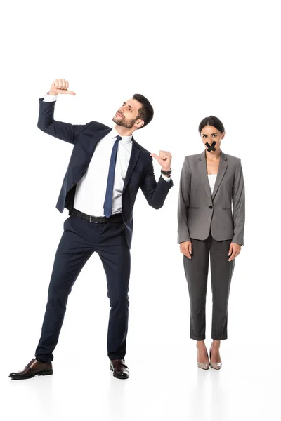 Handsome businessman in formal wear pointing with thumbs at himself near woman with scotch tape on mouth on white — Stock Photo