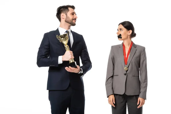 Homme d'affaires tenant trophée d'or et regardant femme d'affaires avec du ruban adhésif sur la bouche isolé sur blanc — Photo de stock