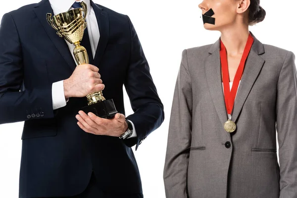 Vista recortada de empresario sosteniendo trofeo cerca de empresaria con cinta adhesiva en la boca y medalla aislada en blanco - foto de stock