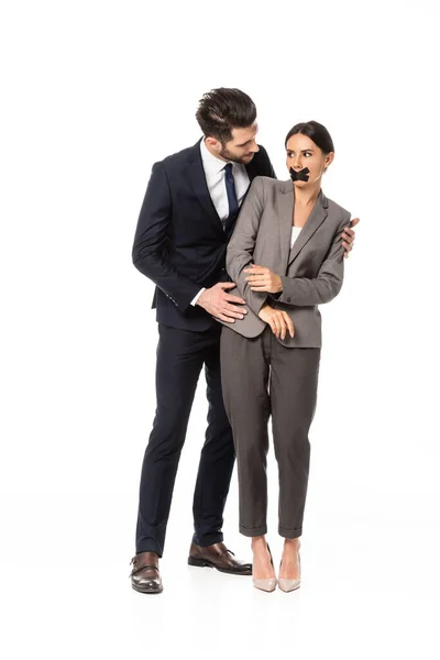 Businessman in formal wear touching businesswoman with duct tape on mouth isolated on white, sexual harassment concept — Stock Photo