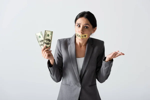 Mujer de negocios emocional con letras de sexismo en cinta adhesiva sosteniendo dólares y mostrando gesto de encogimiento aislado en blanco - foto de stock