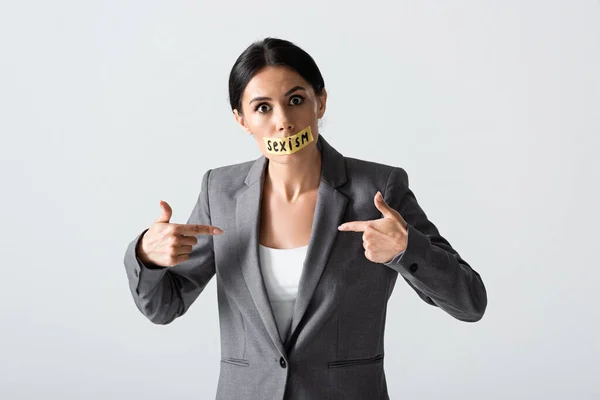 Businesswoman with sexism lettering on duct tape looking at camera while pointing with fingers at herself isolated on white — Stock Photo