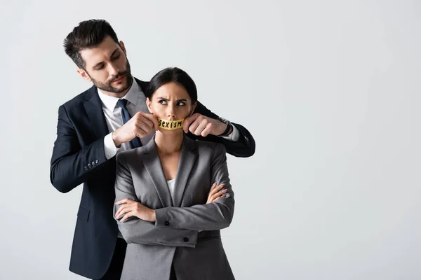 Hombre de negocios barbudo tocando cinta adhesiva con letras de sexismo en la boca de la mujer de negocios de pie con los brazos cruzados aislados en blanco - foto de stock