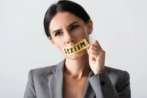 Mujer de negocios tocando cinta adhesiva con letras de sexismo en la boca aislado en blanco - foto de stock