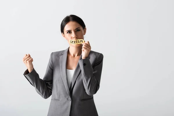 Empresaria mostrando el puño cerrado y quitando cinta adhesiva con letras de sexismo en la boca aislado en blanco - foto de stock