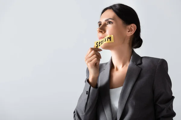 Atractiva mujer de negocios quitando cinta adhesiva con letras de sexismo en la boca aislado en blanco - foto de stock