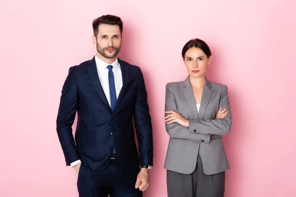 Bell'uomo d'affari in piedi con la mano in tasca vicino a donna d'affari con le braccia incrociate sul rosa, concetto di uguaglianza di genere — Foto stock