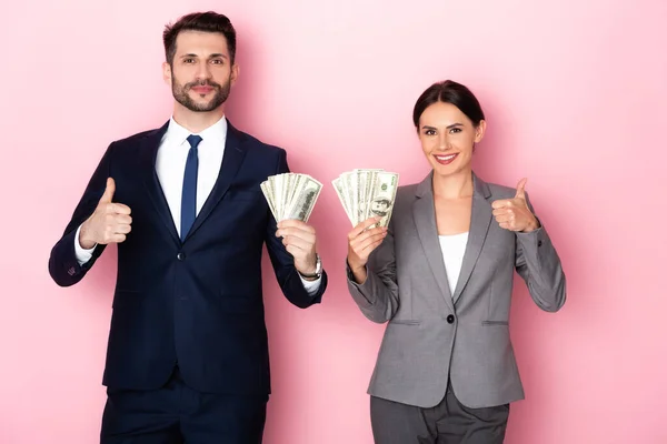 Cheerful businessman and businesswoman holding dollar banknotes and showing thumbs up on pink, gender equality concept — Stock Photo