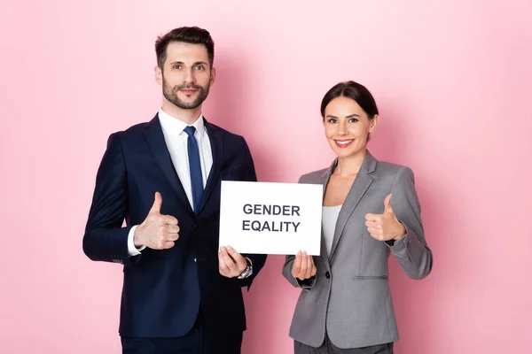 Bel homme d'affaires et femme d'affaires tenant une pancarte avec des lettres d'égalité des sexes et montrant pouces vers le haut sur rose — Photo de stock