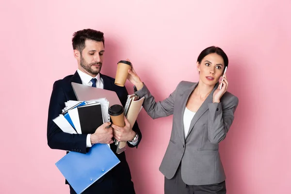 Mujer de negocios hablando en el teléfono inteligente cerca de hombre de negocios sosteniendo vasos de papel, portátil, carpetas y cuadernos en rosa - foto de stock