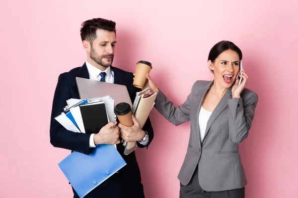 Mujer de negocios enojada hablando en un teléfono inteligente cerca de hombre de negocios sosteniendo vasos de papel, portátil, carpetas y cuadernos en rosa - foto de stock