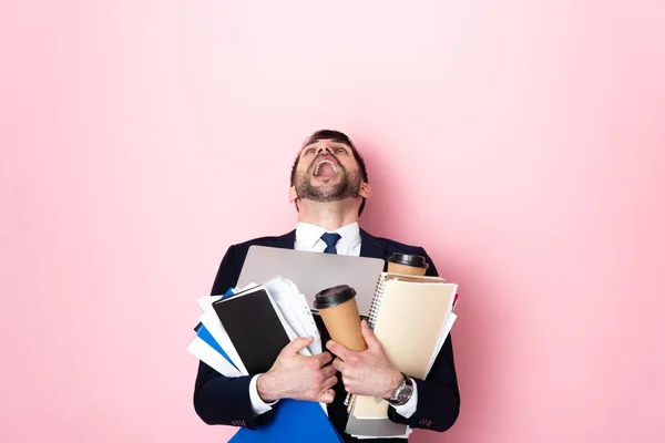 Hombre de negocios barbudo gritando mientras sostiene vasos de papel, portátil, carpetas y cuadernos en rosa - foto de stock