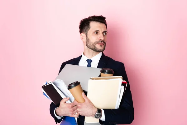 Bel homme d'affaires tenant des tasses en papier, ordinateur portable, dossiers et cahiers sur rose — Photo de stock