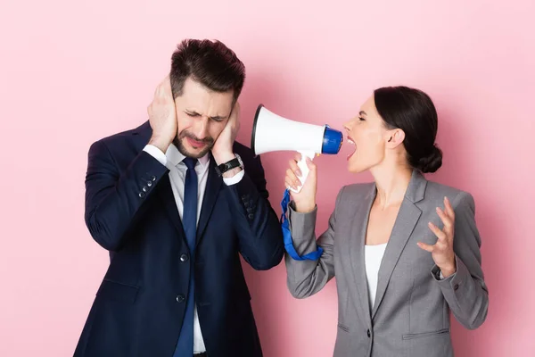 Mujer emocional gritando en megáfono cerca de hombre de negocios barbudo cubriendo las orejas en rosa - foto de stock