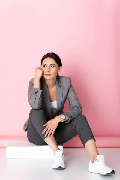 Mujer de negocios pensativa en traje y zapatillas blancas mirando hacia otro lado mientras está sentado en rosa - foto de stock