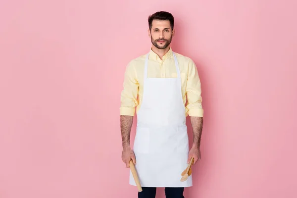 Handsome bearded man holding rolling pin and wooden spoon on pink — Stock Photo