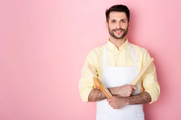 Happy bearded man in apron holding wooden rolling pin and spoon on pink — Stock Photo