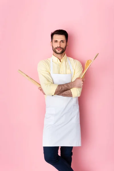 Handsome man in apron holding wooden rolling pin and spoon on pink — Stock Photo