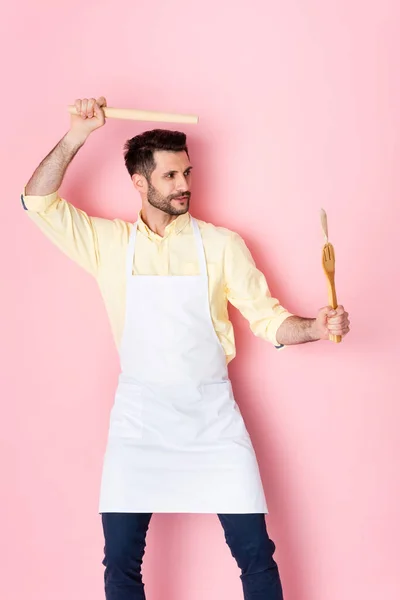 Handsome man in apron holding wooden rolling pin above head on pink — Stock Photo