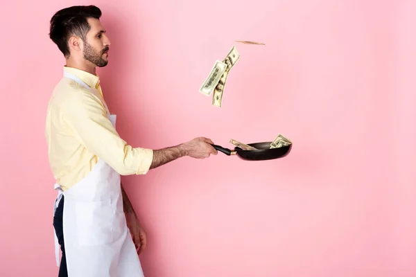Вид сбоку босоногого мужчины в фартуке, держащего в руках сковороду с деньгами на розовом фоне — стоковое фото