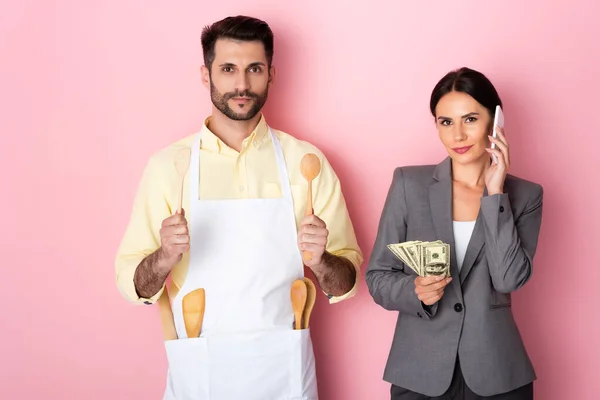 Hombre guapo en delantal sosteniendo cuchara de madera y espátula cerca de la mujer de negocios con dinero hablando en el teléfono inteligente en rosa - foto de stock