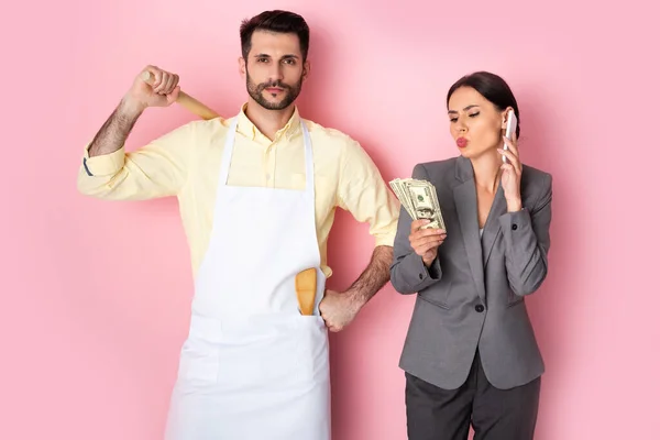 Handsome man in apron holding wooden rolling pin near businesswoman with money talking on smartphone on pink — Stock Photo