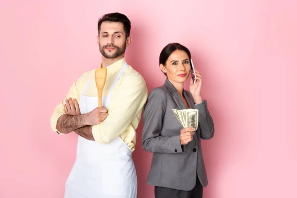 Bel homme dans tablier tenant spatule en bois près d'une femme d'affaires avec de l'argent parlant sur smartphone sur rose — Photo de stock