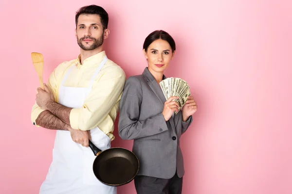 Bel homme dans tablier tenant spatule en bois et poêle près d'une femme d'affaires avec de l'argent sur rose — Photo de stock