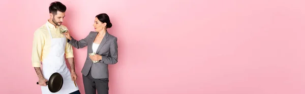 Заголовок веб-сайту бізнес-леді, що кладе гроші в кишеню красивого чоловіка в фартух, тримає сковороду на рожевому — стокове фото