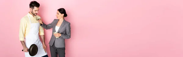 Website header of businesswoman putting money in pocket of handsome man in apron holding frying pan on pink — Stock Photo