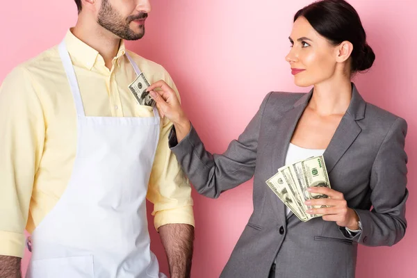 Businesswoman putting money in pocket of bearded man in apron on pink — Stock Photo