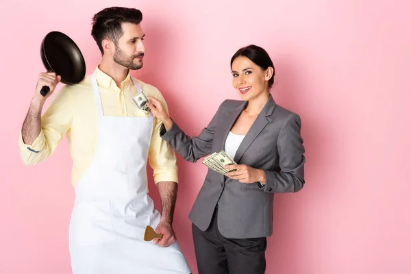 Empresária feliz colocando dinheiro no bolso de homem bonito em avental segurando frigideira e espátula em rosa — Fotografia de Stock