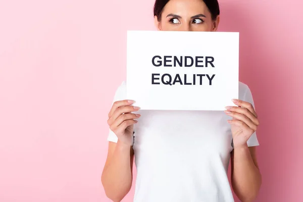 Mujer en camiseta blanca cubriendo la cara con letras de igualdad de género en el cartel y mirando hacia otro lado en rosa - foto de stock