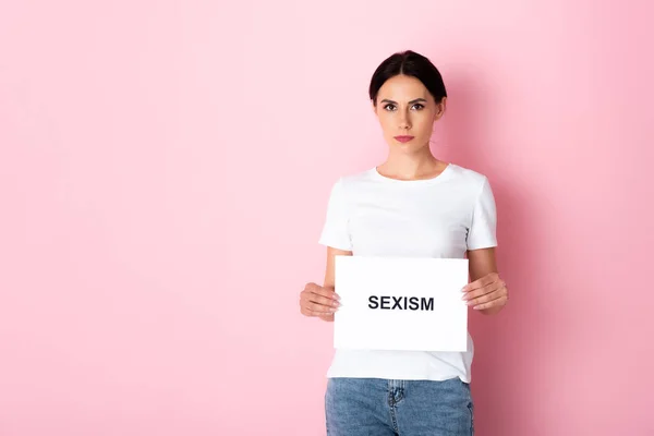 Jolie femme en t-shirt blanc tenant placard avec lettrage sexisme sur rose — Photo de stock