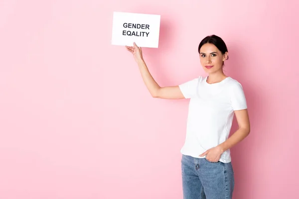 Mulher feliz em t-shirt branca segurando cartaz com letras igualdade de gênero e de pé com a mão no bolso em rosa — Fotografia de Stock