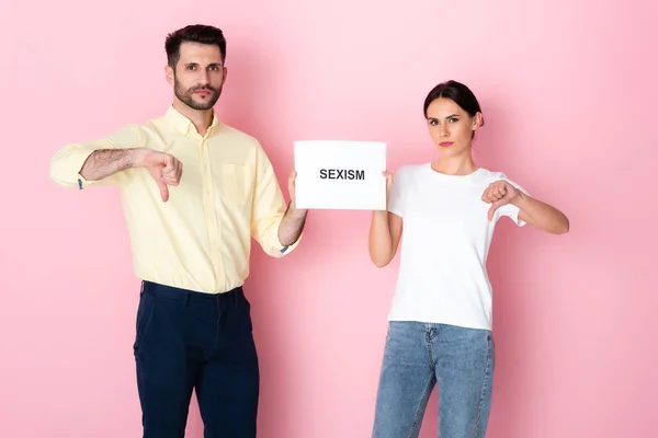 Mann und Frau in weißem T-Shirt halten Plakat mit Sexismus-Schriftzug hoch und zeigen Daumen nach unten auf rosa — Stockfoto
