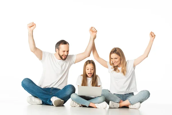 Excited parents sitting on floor near happy daughter with laptop isolated on white — Stock Photo