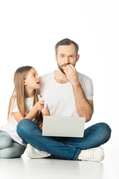 Scioccato padre e figlia seduti sul pavimento con computer portatile isolato su bianco — Foto stock